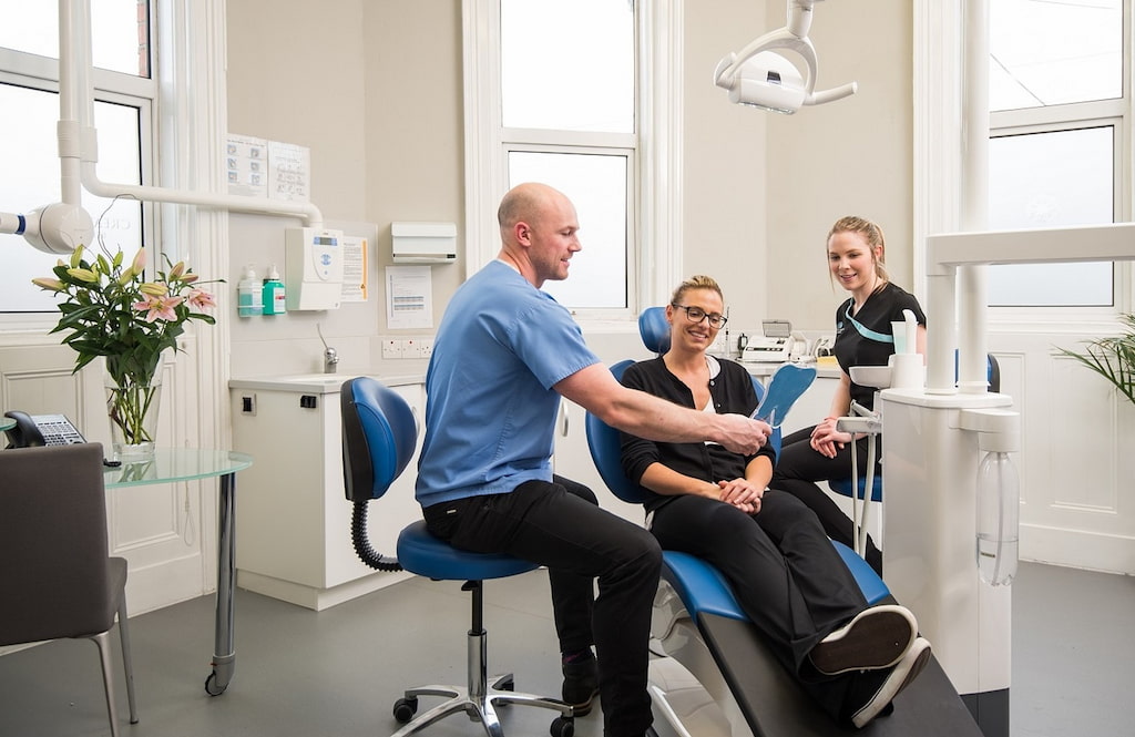 Foto på en leende kvinna i tandläkarstol i mottagningsrummet, med sköterska och tandläkare på varsin sida.