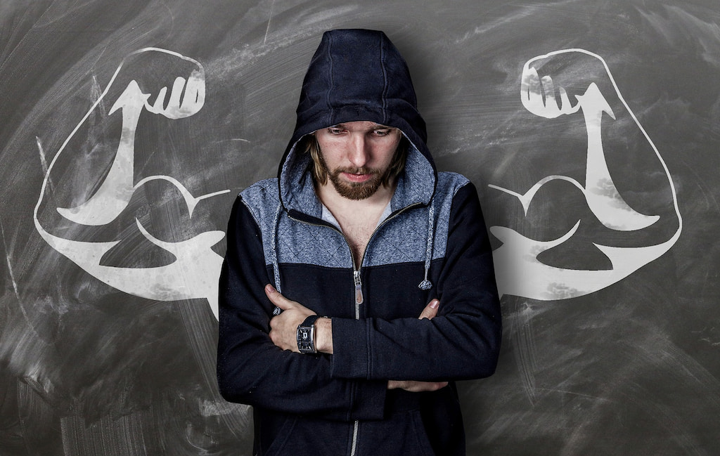 En man med hoodie står med armarna i kort med ryggen mot svarta tavlan, vilken har uppritade muskulösa armar uppsträckta bakom mannen.